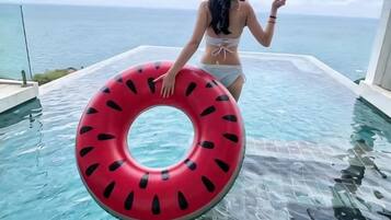 Mulher de biquíni branco segura um flutuador em formato de argola vermelha em uma piscina infinita transparente, com vista para o oceano.