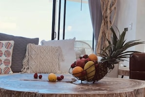 Een ronde houten tafel met een fruitschaal met appels, sinaasappels en een ananas, een geweven stoel en uitzicht op het landschap.