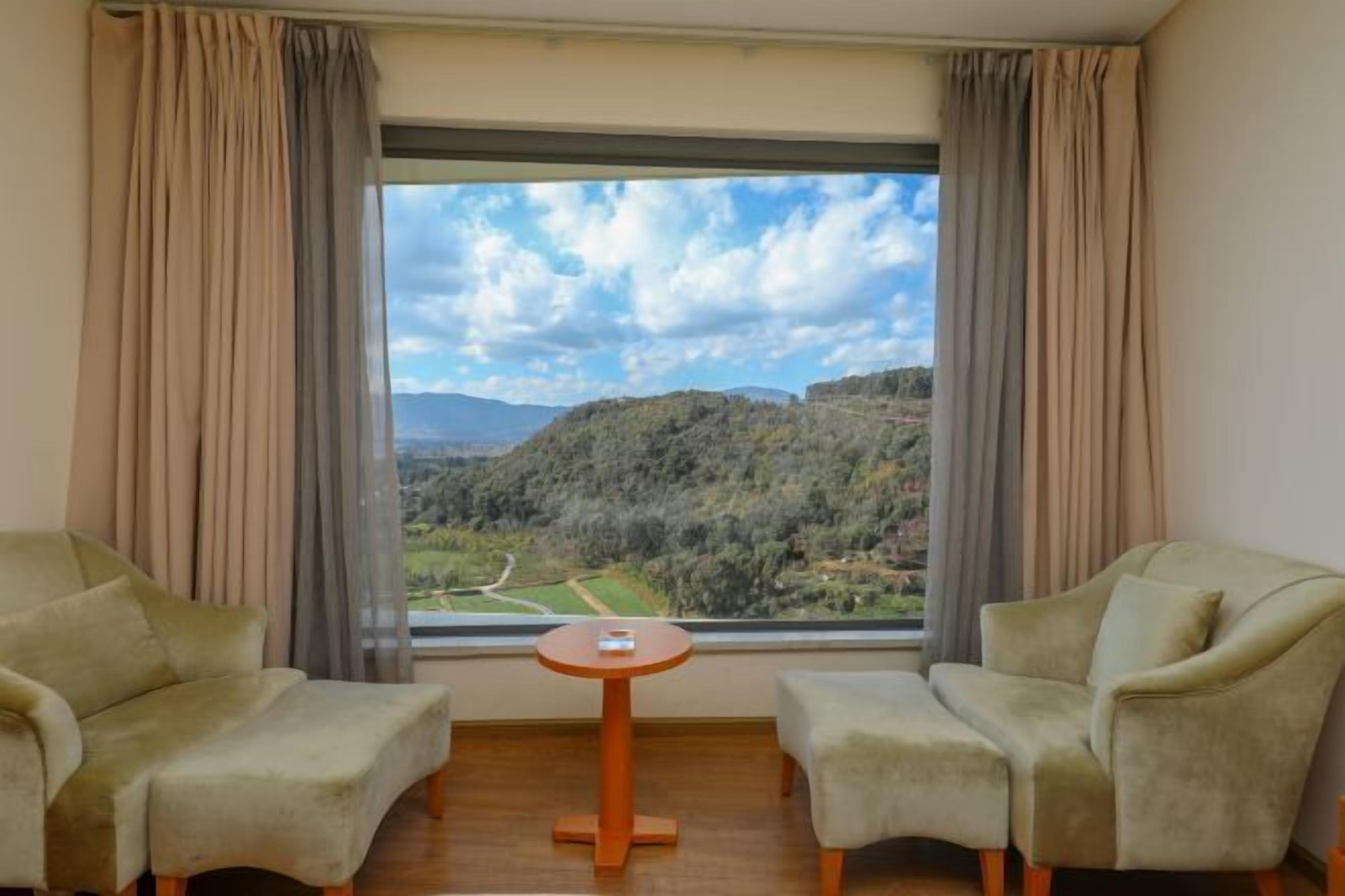 A room with two armchairs and a small table, a large window with curtains, and a view of a hilly landscape.