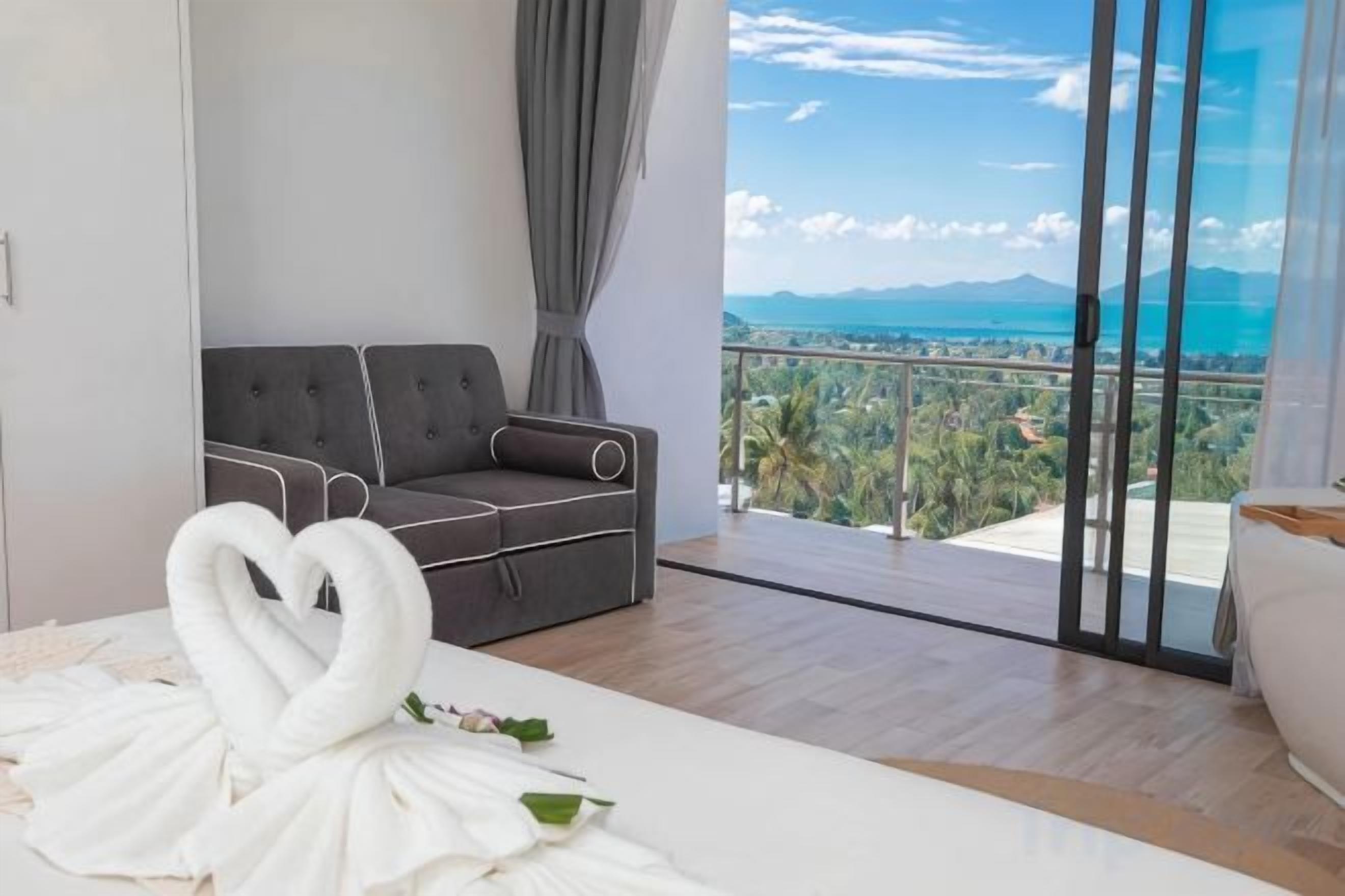 Habitación de hotel moderna con sofá, una toalla dispuesta en forma de corazón y un balcón con vistas al mar y a las montañas.