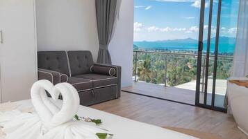 Habitación de hotel moderna con sofá, una toalla dispuesta en forma de corazón y un balcón con vistas al mar y a las montañas.
