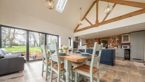 A spacious dining area with a wooden table and chairs, a kitchen in the background, and a view of the outdoors. - Waterside Barn, Binham, Norfolk (Binham)