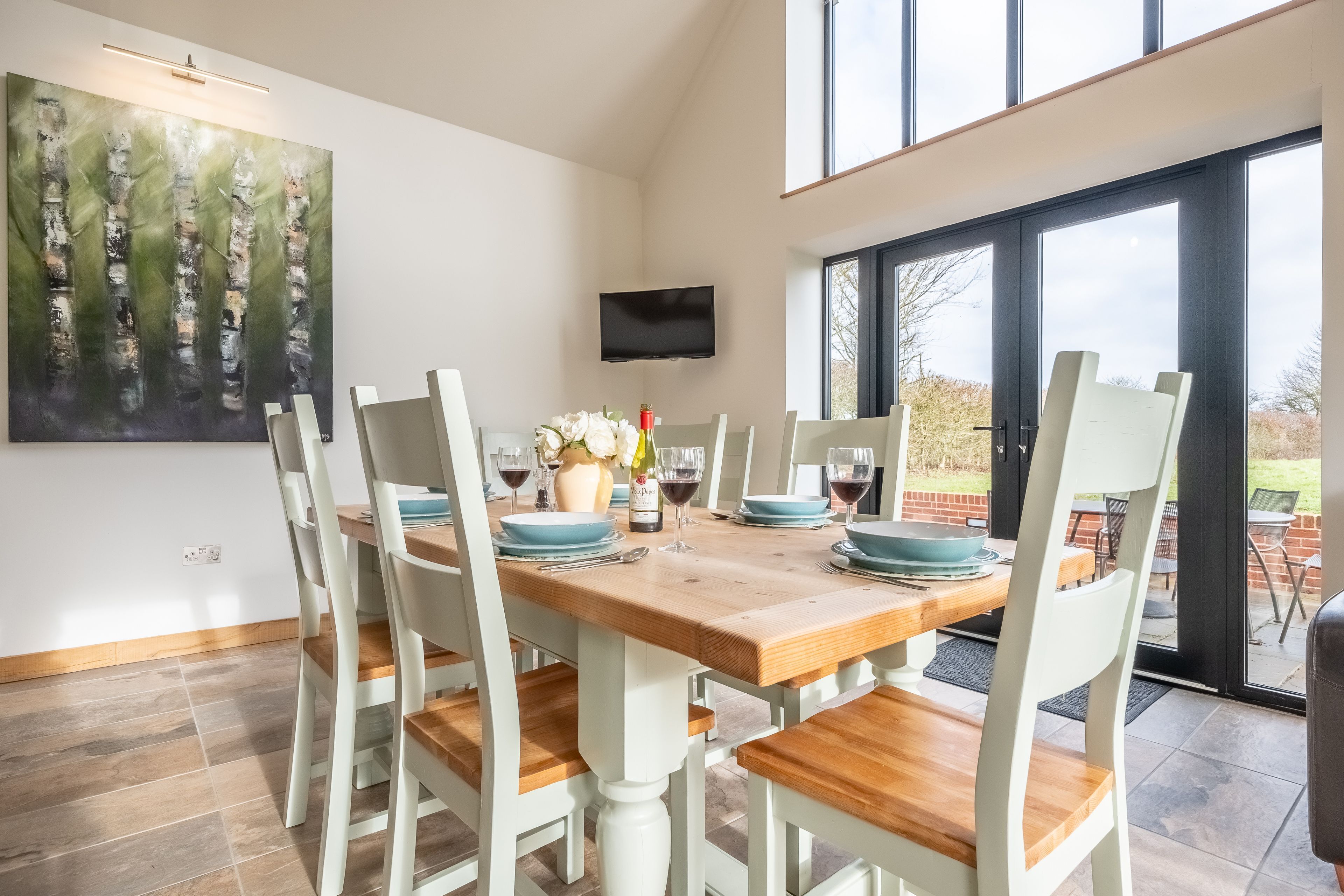 A dining area with a wooden table set for six, white chairs, and a large painting on the wall.