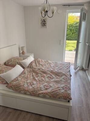 Une chambre à coucher comprenant un lit, une table de chevet, une lampe, un lustre, une porte donnant sur un balcon et un tableau encadré accroché au mur.