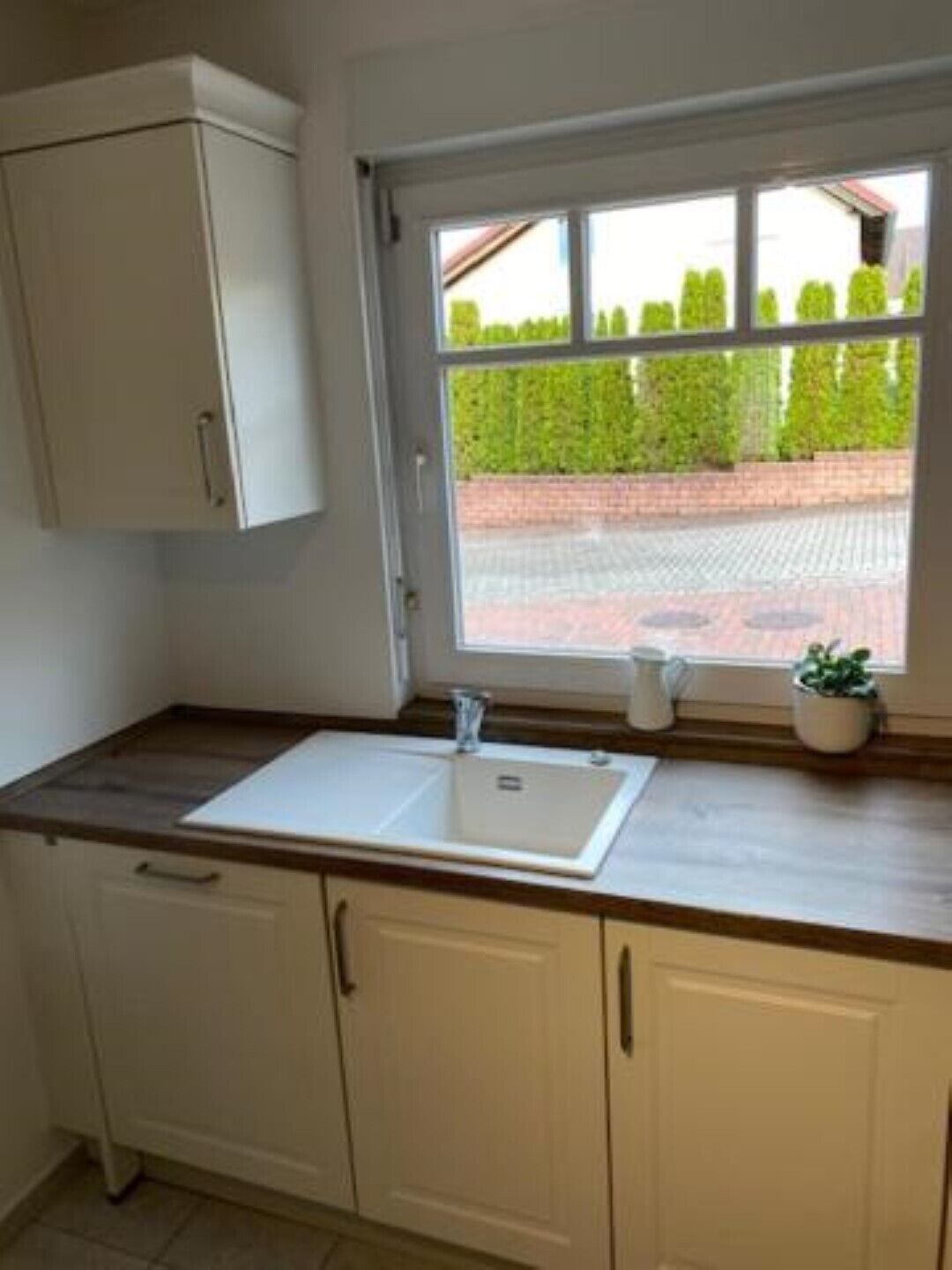 A kitchen with a white sink, cabinets, and a window with a view of a brick wall and greenery.