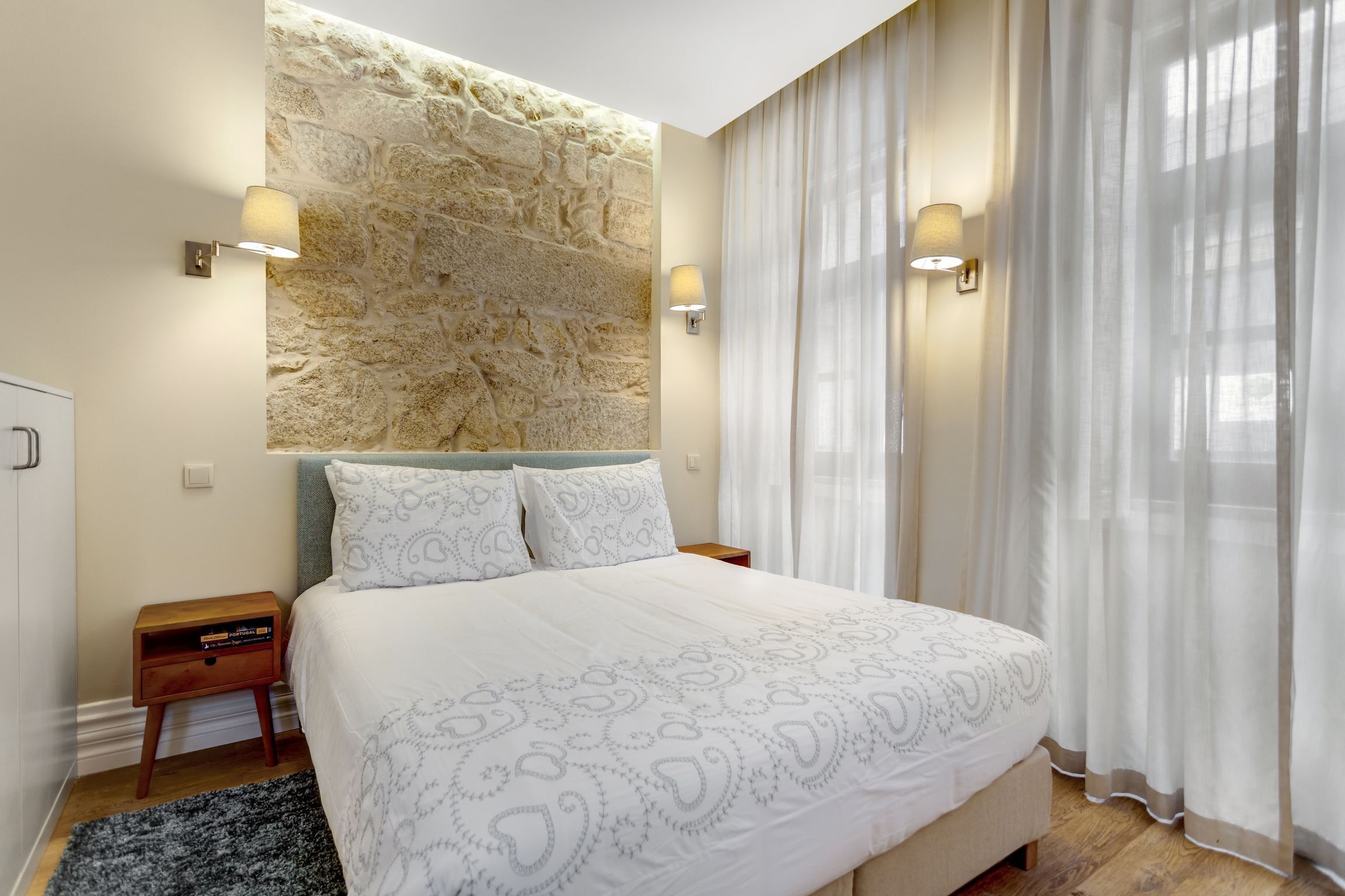 A bedroom with a large bed, stone wall, two bedside lamps, and sheer curtains.