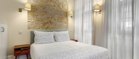 A bedroom with a large bed, stone wall, two bedside lamps, and sheer curtains.