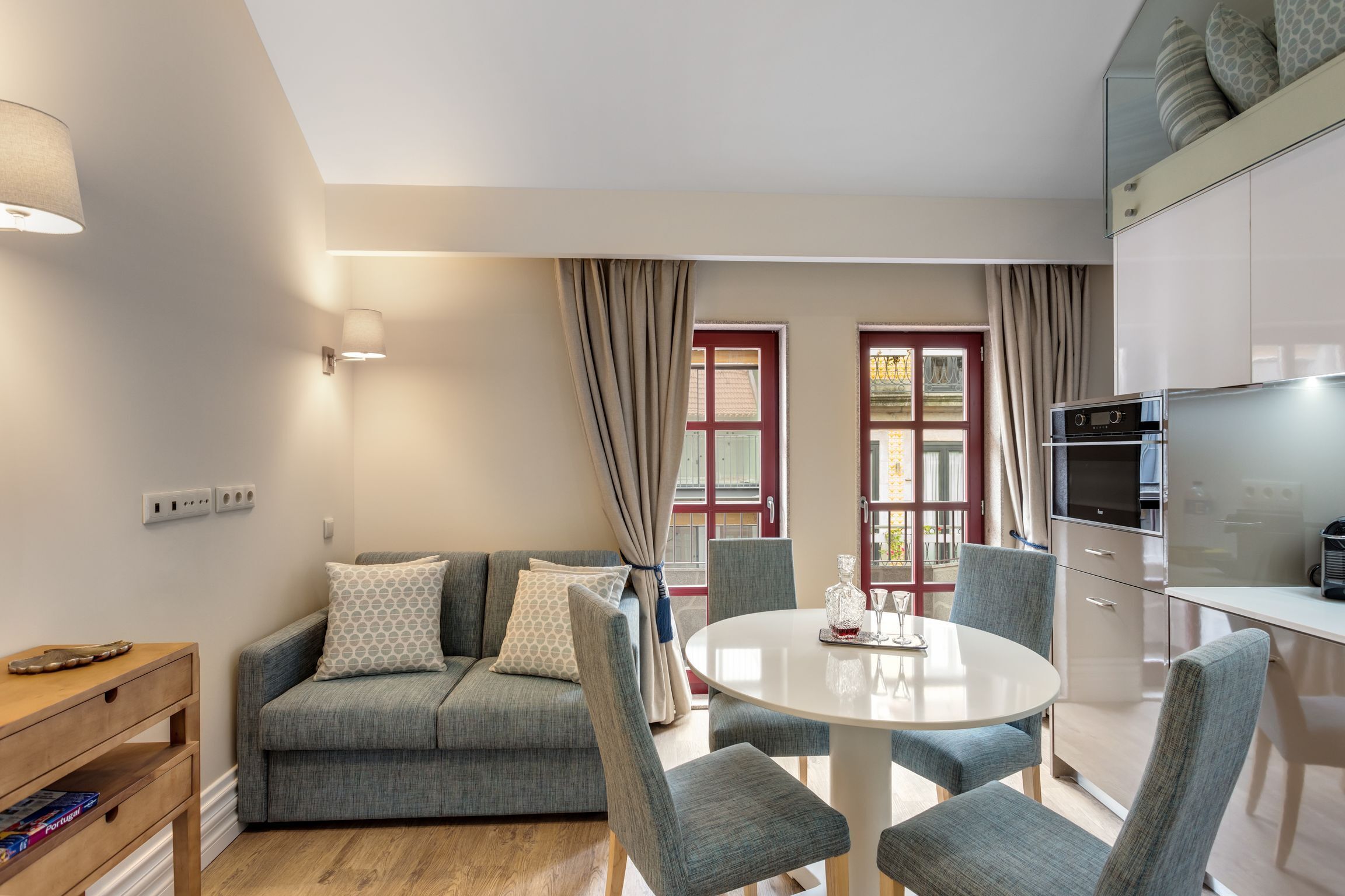 Ein modernes Wohnzimmer mit einem grauen Sofa, einem runden Esstisch und passenden Stühlen. Es gibt eine Küchenzeile mit Kühlschrank und Mikrowelle.