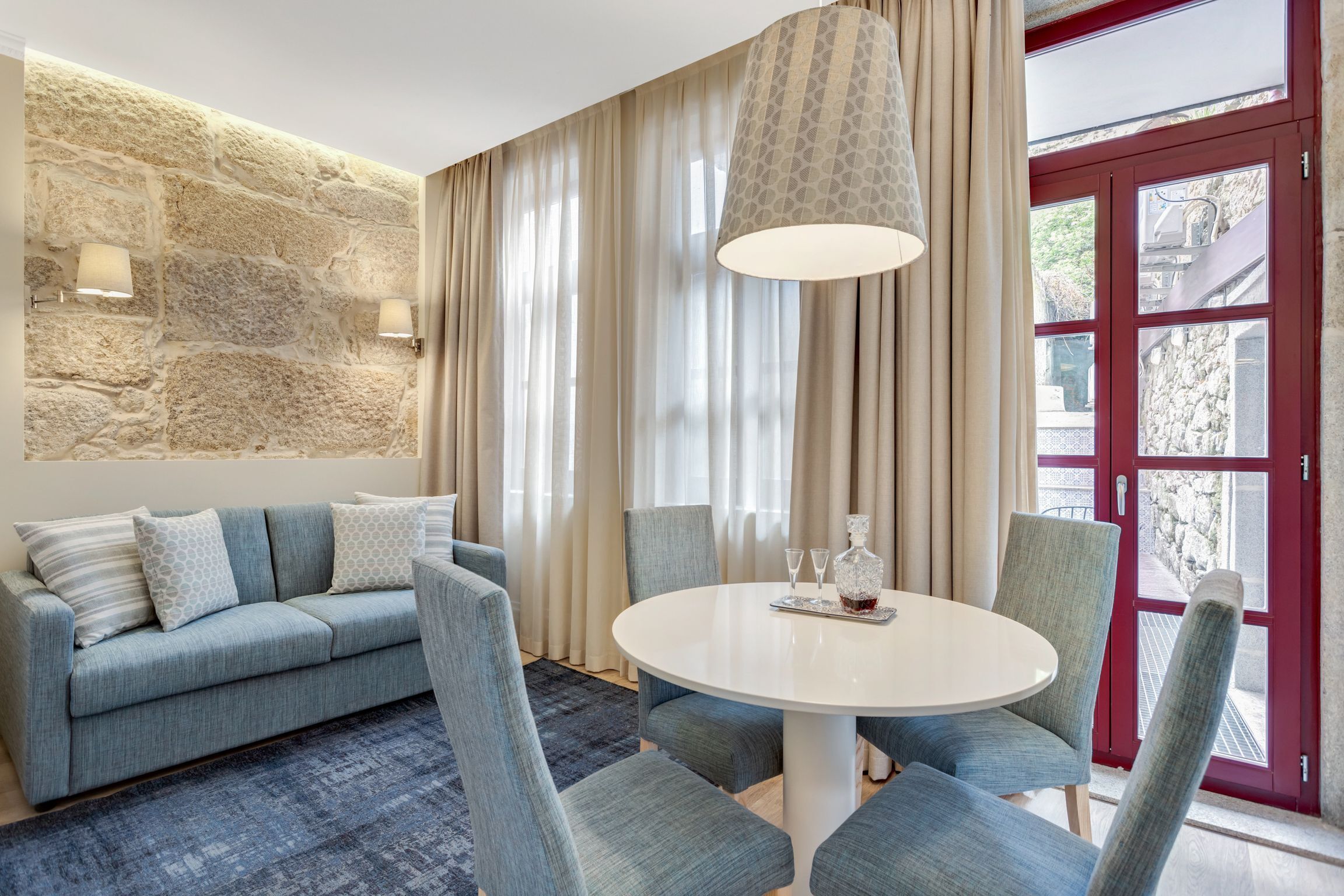 Una habitación con pared de piedra, un sofá azul con cojines estampados, una mesa redonda blanca con sillas y una puerta roja con paneles de vidrio.