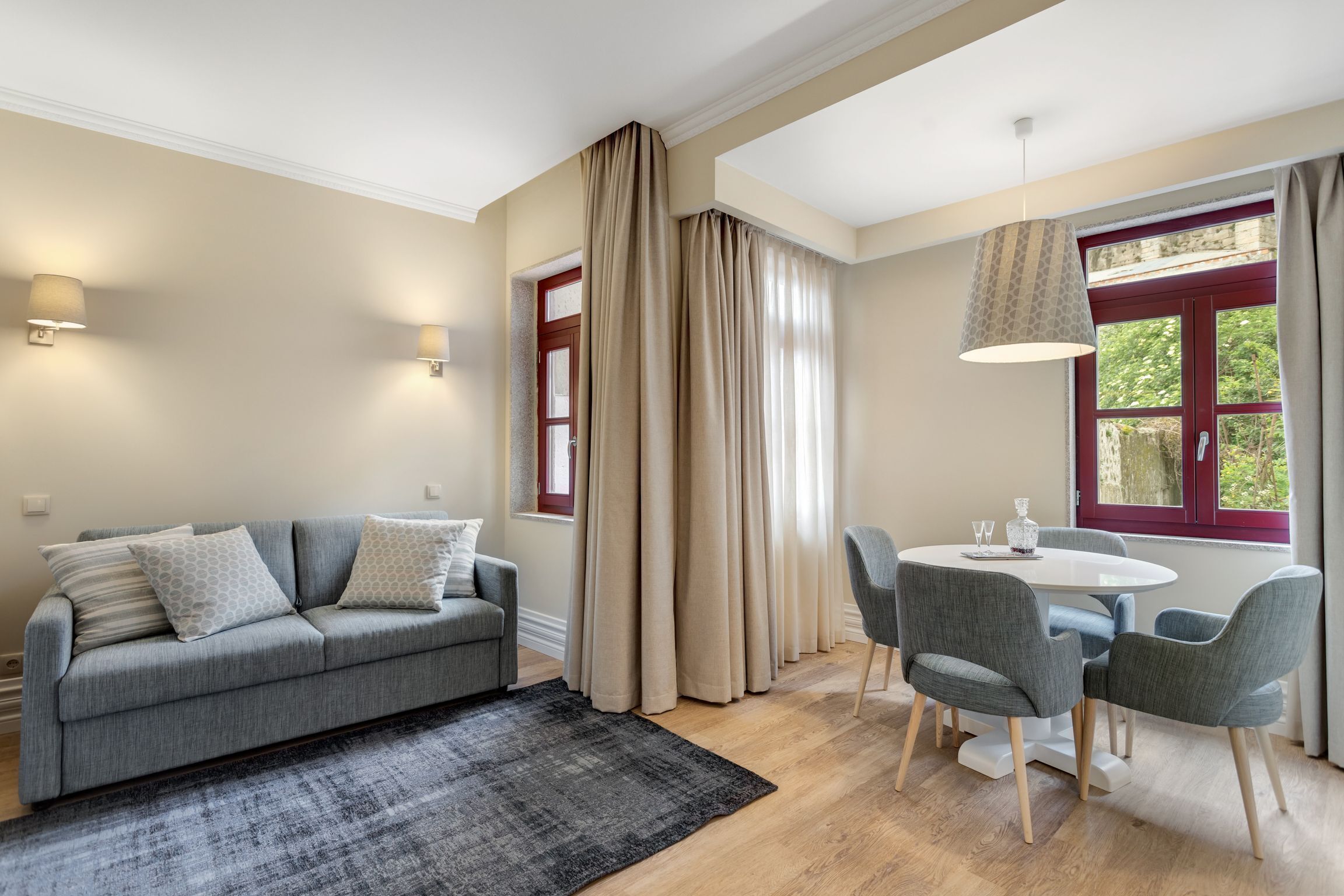 A modern living room with a grey sofa, a round dining table with four chairs, and a large window with curtains.