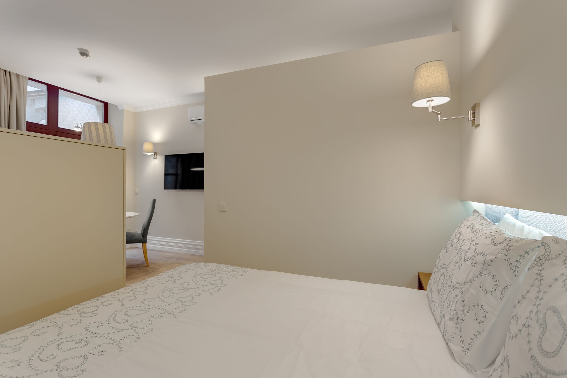 Une chambre à coucher avec un lit, une table de chevet, une lampe fixée au mur et une fenêtre avec des rideaux.