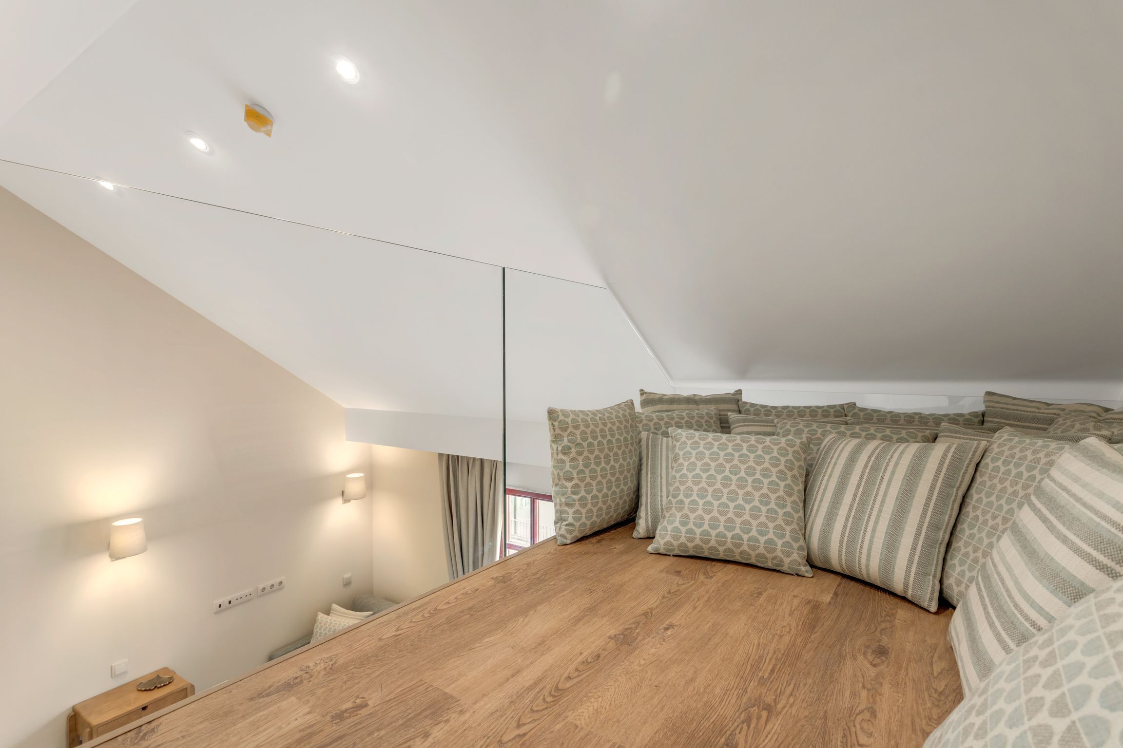 A modern bedroom with a sloped ceiling, wooden flooring, and a bed with patterned pillows.