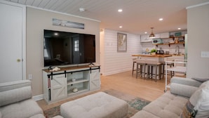 A modern living room with a large flat-screen TV, a wooden coffee table, and two grey sofas. A kitchen with white cabinets and a bar area is visible in the background. - New Cottage: 4th row & fully fenced back yard! (Hilton Head Island)