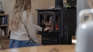 A person sitting in front of a wood-burning stove in a room with a bookshelf and a window. - Cozy Cottage in Locust Lake w/ Seasonal Lake Views (pocono Lake)