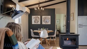 A cozy living room with a person reading, a dining area with a round table, and a wood-burning stove. - Cozy Cottage in Locust Lake w/ Seasonal Lake Views (pocono Lake)
