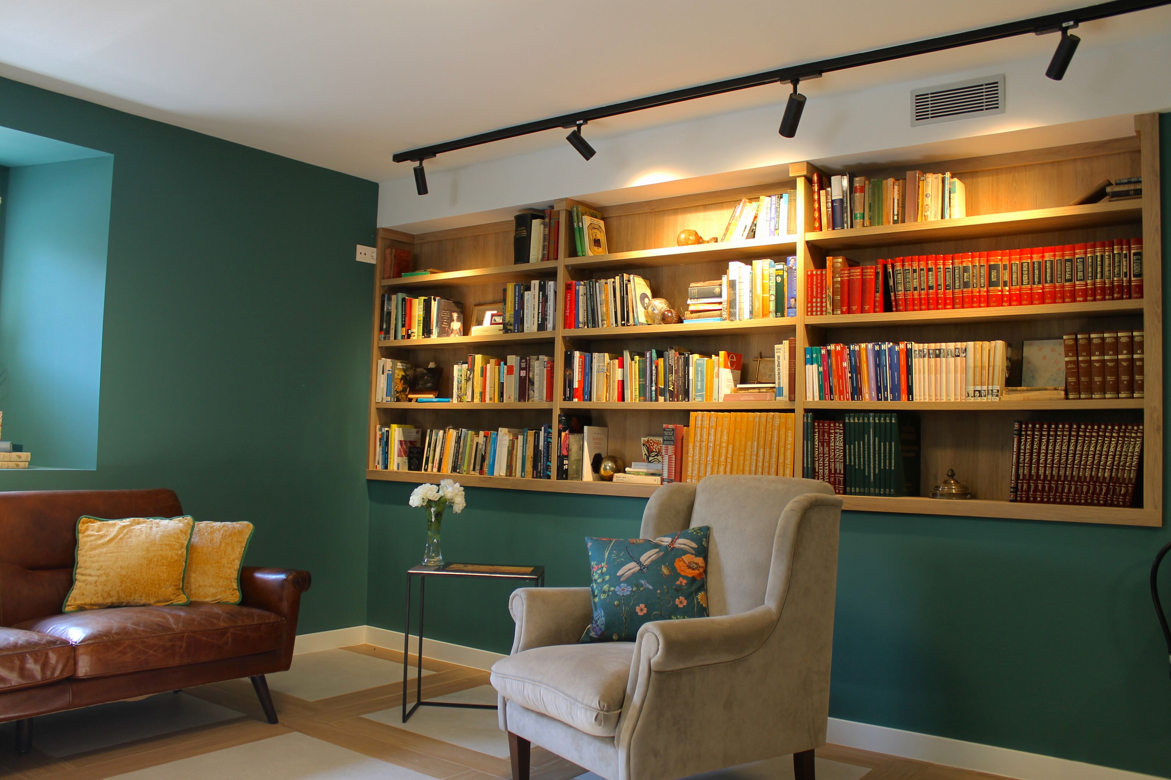 Una sala de estar acogedora con un sofá de cuero marrón, un sillón color crema y una estantería llena de libros.