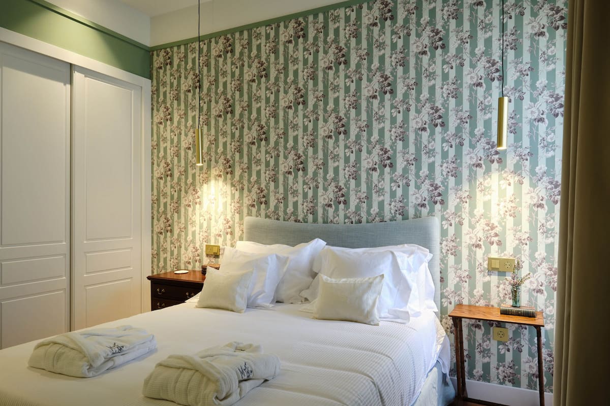 Un lit bien fait, avec du linge de lit blanc et des oreillers, une table de chevet avec une lampe, et un papier peint à motifs floraux.