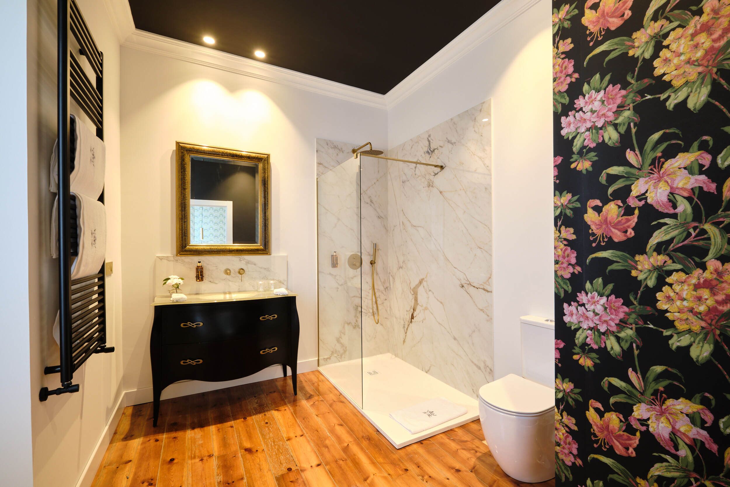Ein Badezimmer mit Holzboden, einem schwarzen Waschtisch mit goldfarbenen Griffen, einem weißen Waschbecken, einer Glasdusche und einer Toilette.