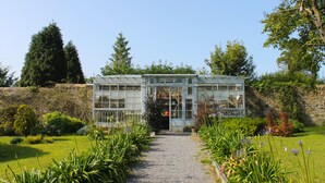 A greenhouse with a gravel path leading to it, surrounded by various plants and trees.