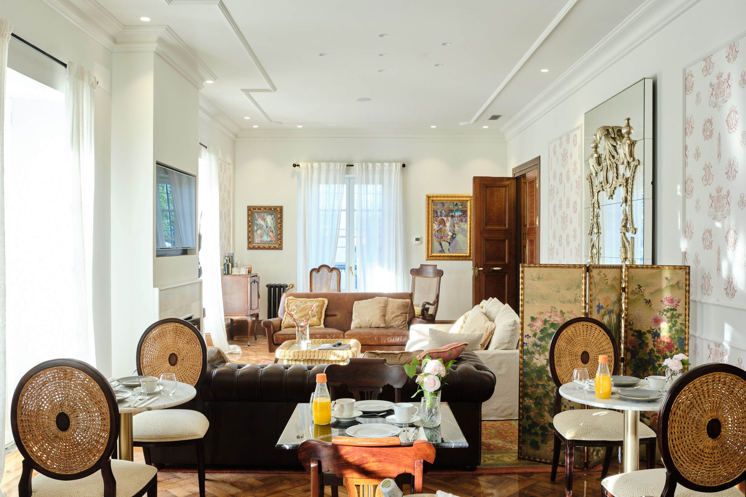 Un salón amplio con un sofá negro, dos sillones de mimbre, una mesa de comedor con un jarrón de flores y un cuadro grande en la pared.