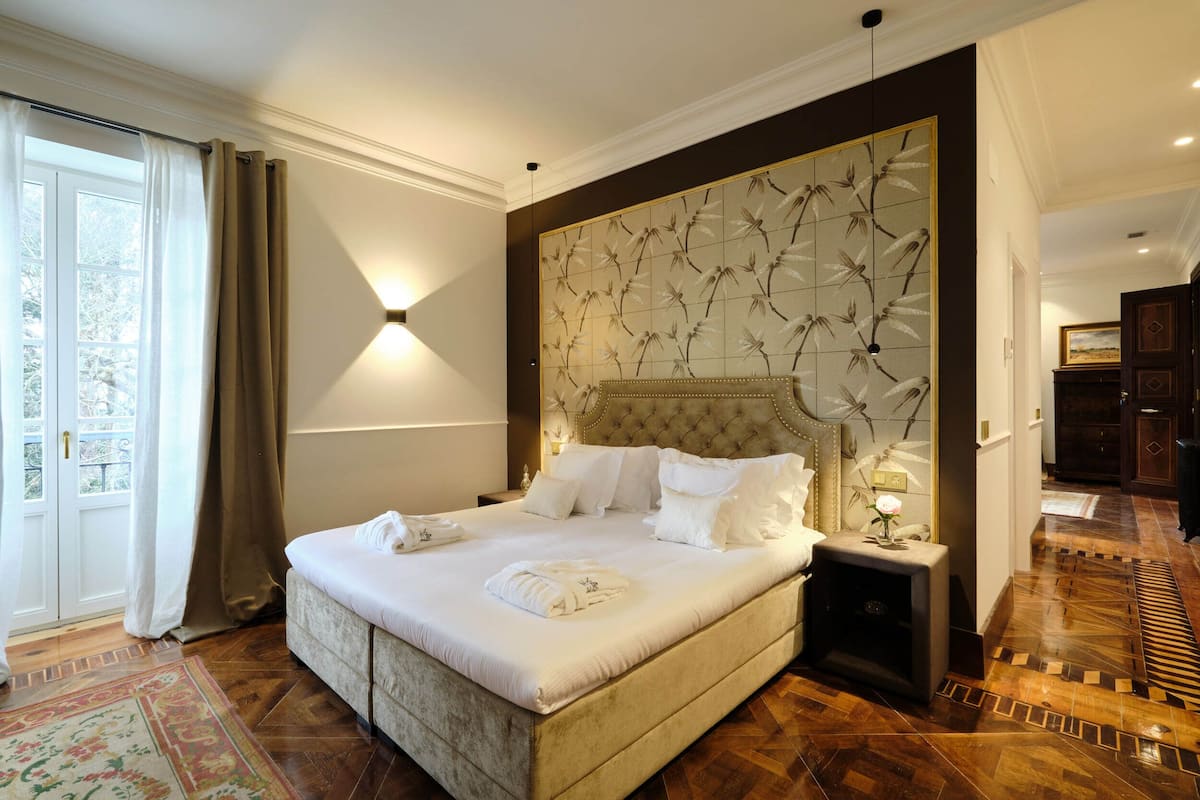 Un grand lit avec du linge de lit blanc et une tête de lit ornée d’un motif floral, une table de chevet avec une lampe, et une vue sur un plancher en bois et une porte.