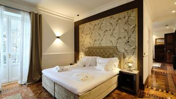 A large bed with white linens and a headboard with a floral pattern, a bedside table with a lamp, and a view of a wooden floor and a door.