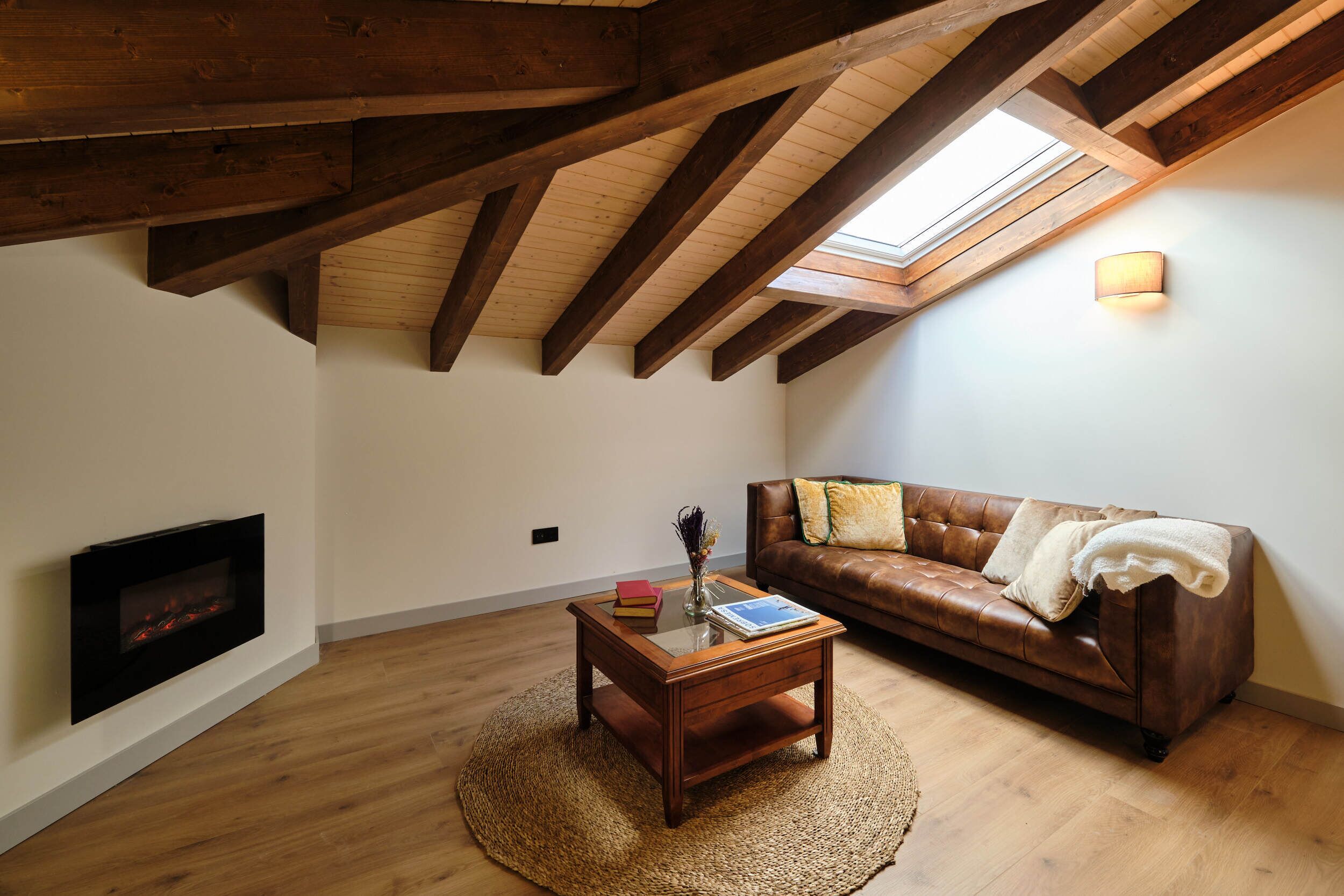 Una habitación estilo loft con vigas de madera a la vista, chimenea, sofá de cuero y tragaluz.