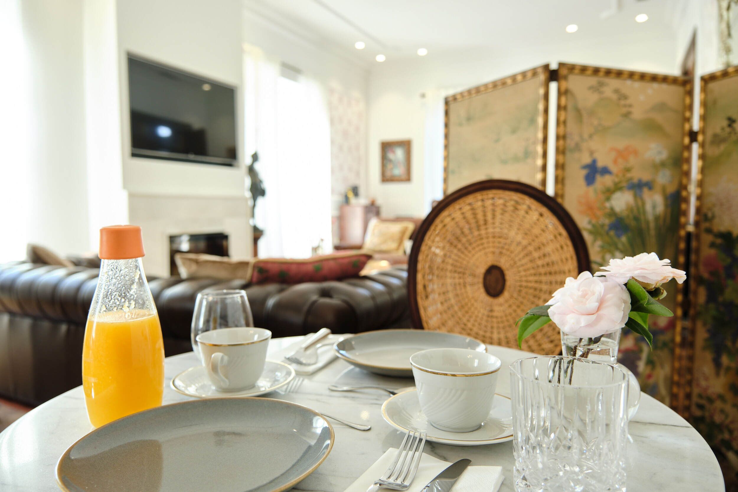 A dining table set with plates, cups, and a glass of orange juice, with a sofa and decorative panels in the background.