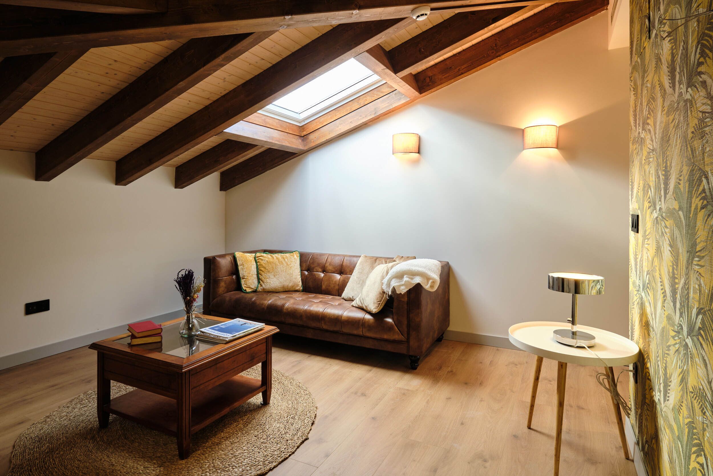Un dormitorio estilo loft acogedor con techo inclinado, un sofá de cuero, una mesita redonda y una lámpara de pared.