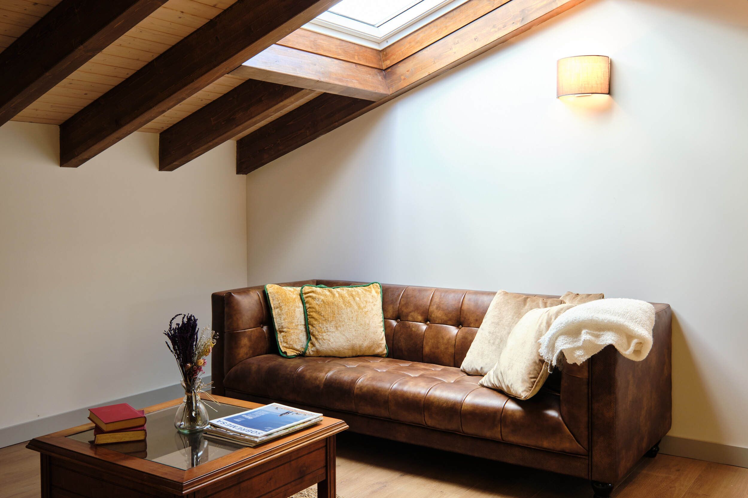 Una habitación acogedora con un sofá de cuero marrón, una mesa de centro de madera con libros, un jarrón con flores secas y un tragaluz.