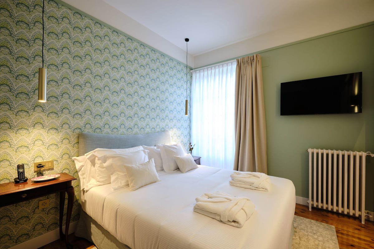 Un lit bien fait, avec du linge de lit blanc, une table de chevet avec un téléphone et une assiette, un téléviseur fixé au mur et un radiateur.