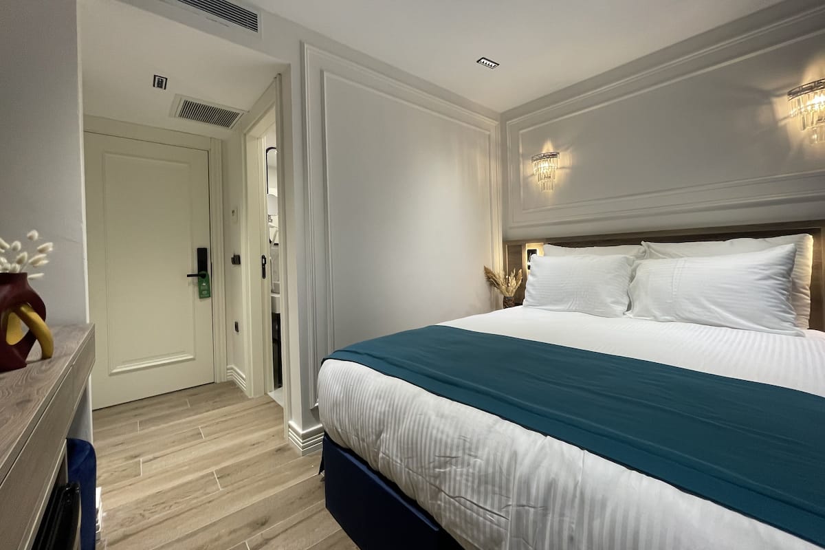 Een hotelkamer met een groot bed, een houten vloer, een deur naar een badkamer en een wandlamp.