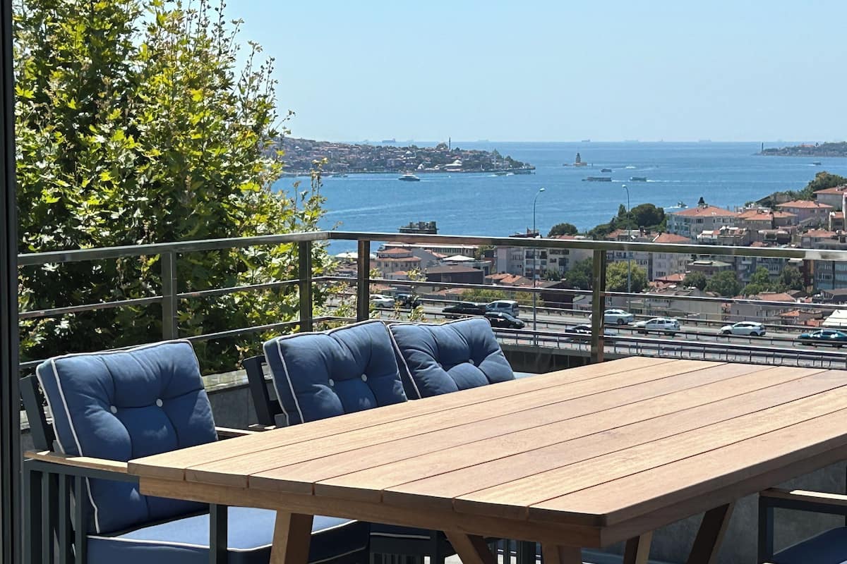 Een houten tafel en blauwe stoelen op een balkon met uitzicht op een kustplaats, met gebouwen en boten.
