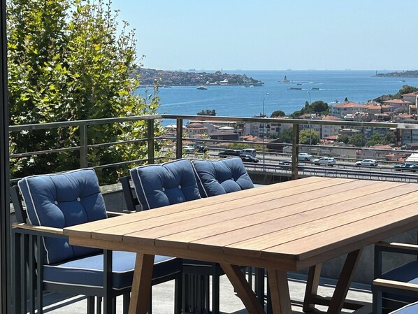 A wooden table and blue chairs on a balcony overlooking a coastal city with visible buildings and boats. - Mona Bosphorus  (Istanbul)