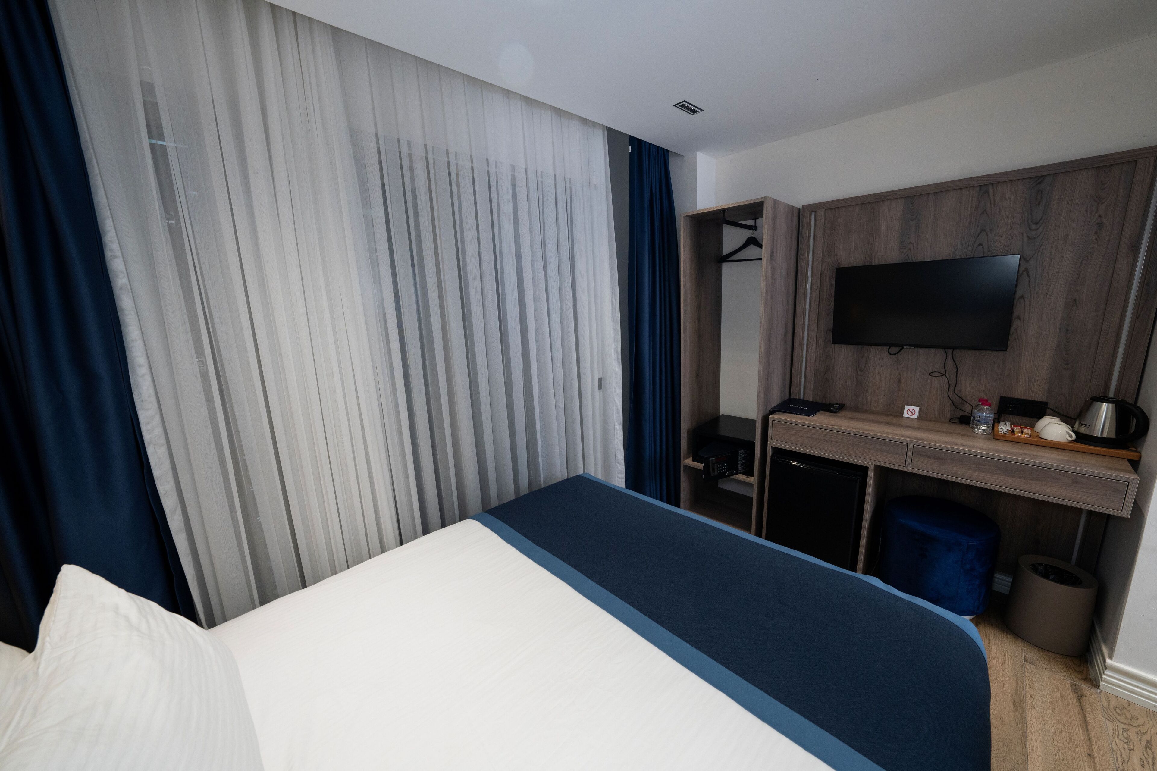 A hotel room with a bed, a desk with a flat-screen TV, a wardrobe, and a trash bin.
