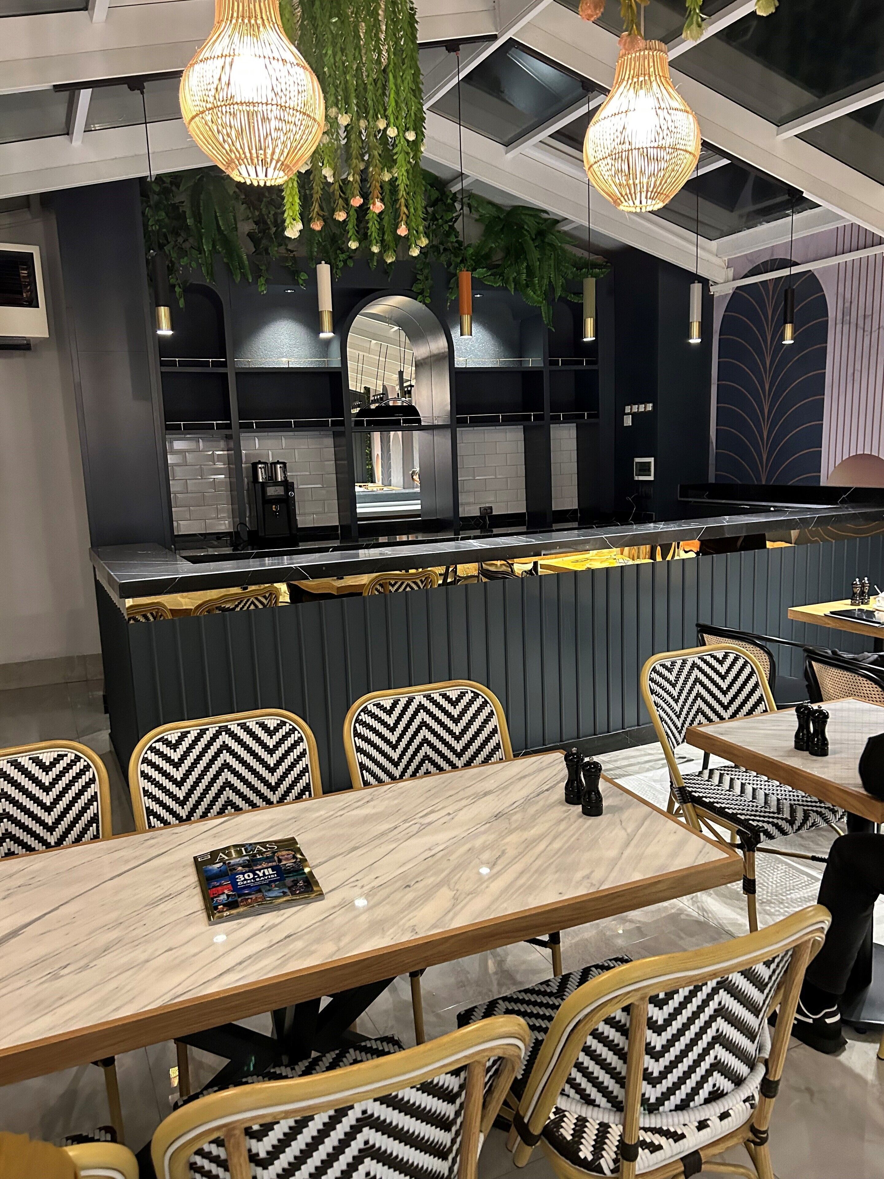 Un intérieur de restaurant moderne avec un bar central, des chaises à motifs noir et blanc, des luminaires suspendus et une cuisine ouverte.