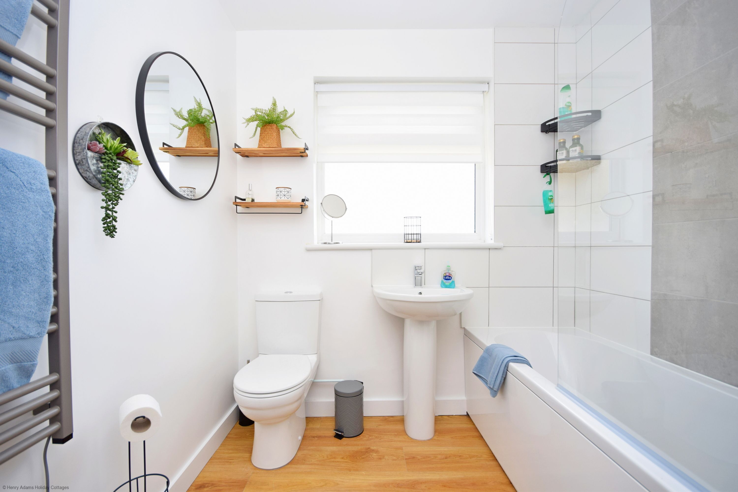 A modern bathroom with a toilet, sink, and bathtub. There are shelves with plants and toiletries.