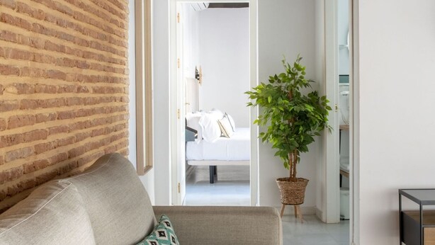 A modern hotel room with a brick wall, a grey sofa, a potted plant, and a bed with white bedding. - Hommyhome Triana Boutique Apartments 004 (Sevilla)
