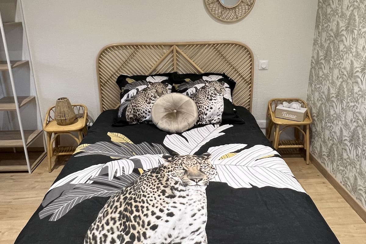 Ein Schlafzimmer mit einem Bett, dessen Bettdecke ein Leopardenmuster aufweist, einem hölzernen Kopfteil und dekorativen Kissen. Es gibt zwei Nachttische mit kleinen Körbchen und einer Taschentuchbox. Die Wand ist weiß, mit einem runden Spiegel und einer Wandtapete mit Muster auf einer Seite.