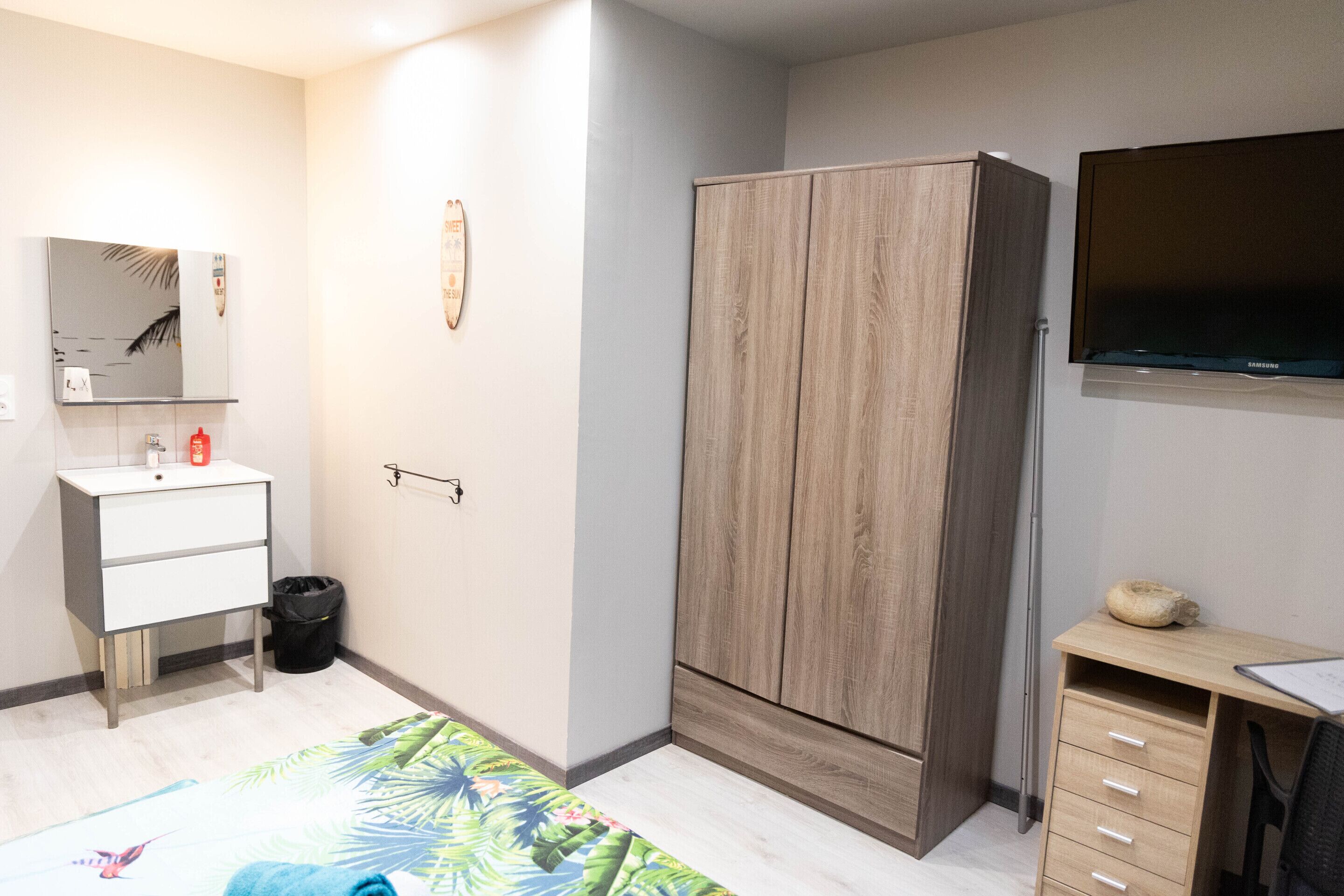 A bedroom with a bed, a wardrobe, a desk, a mirror, and a television.