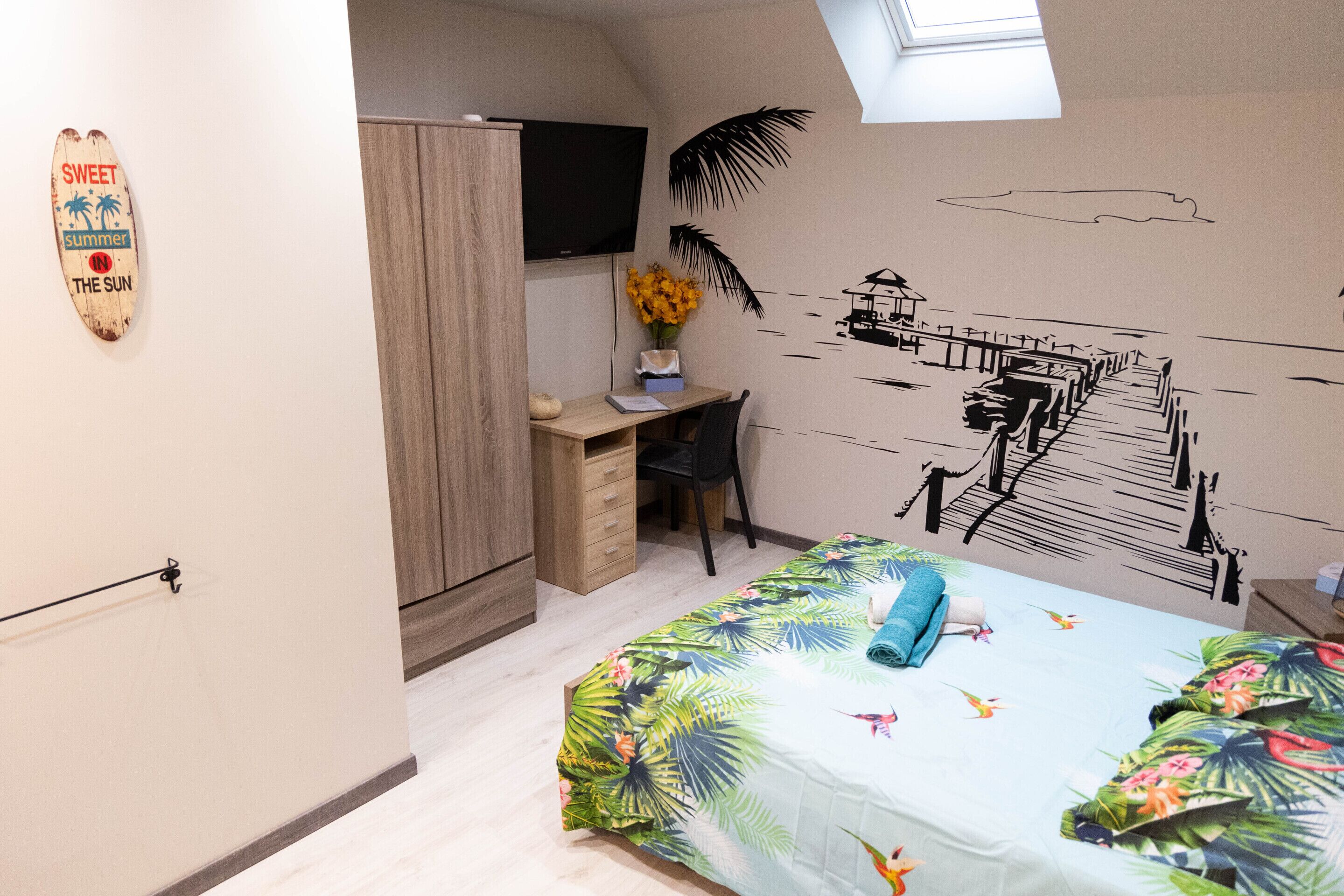 A bedroom with a bed, a desk, a chair, a TV, and a wall with a tropical mural.