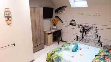 A bedroom with a bed, a desk, a chair, a TV, and a wall with a tropical mural.