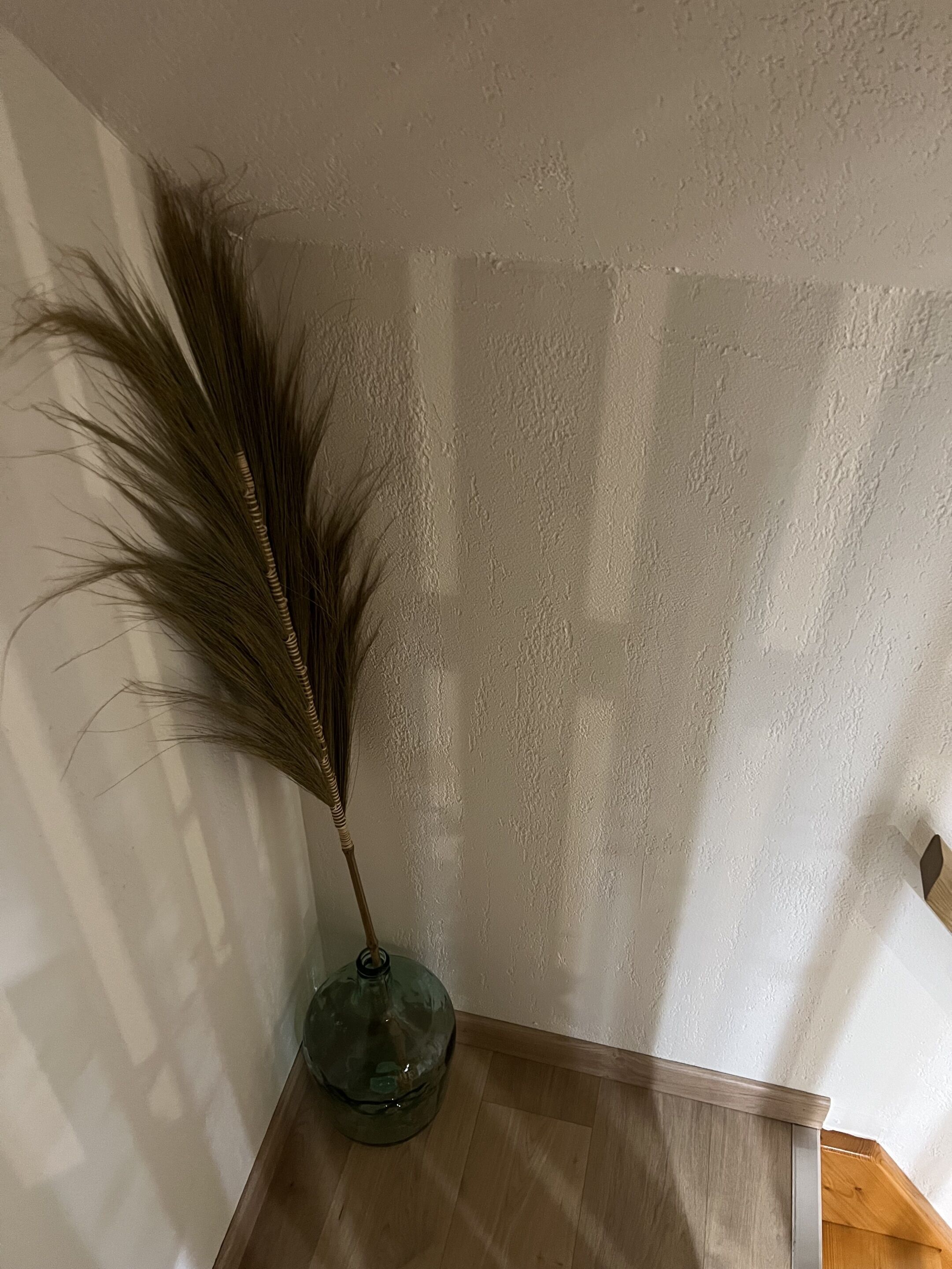 A corner of a room with a wooden floor, a white textured wall, and a green vase with a dried plant.