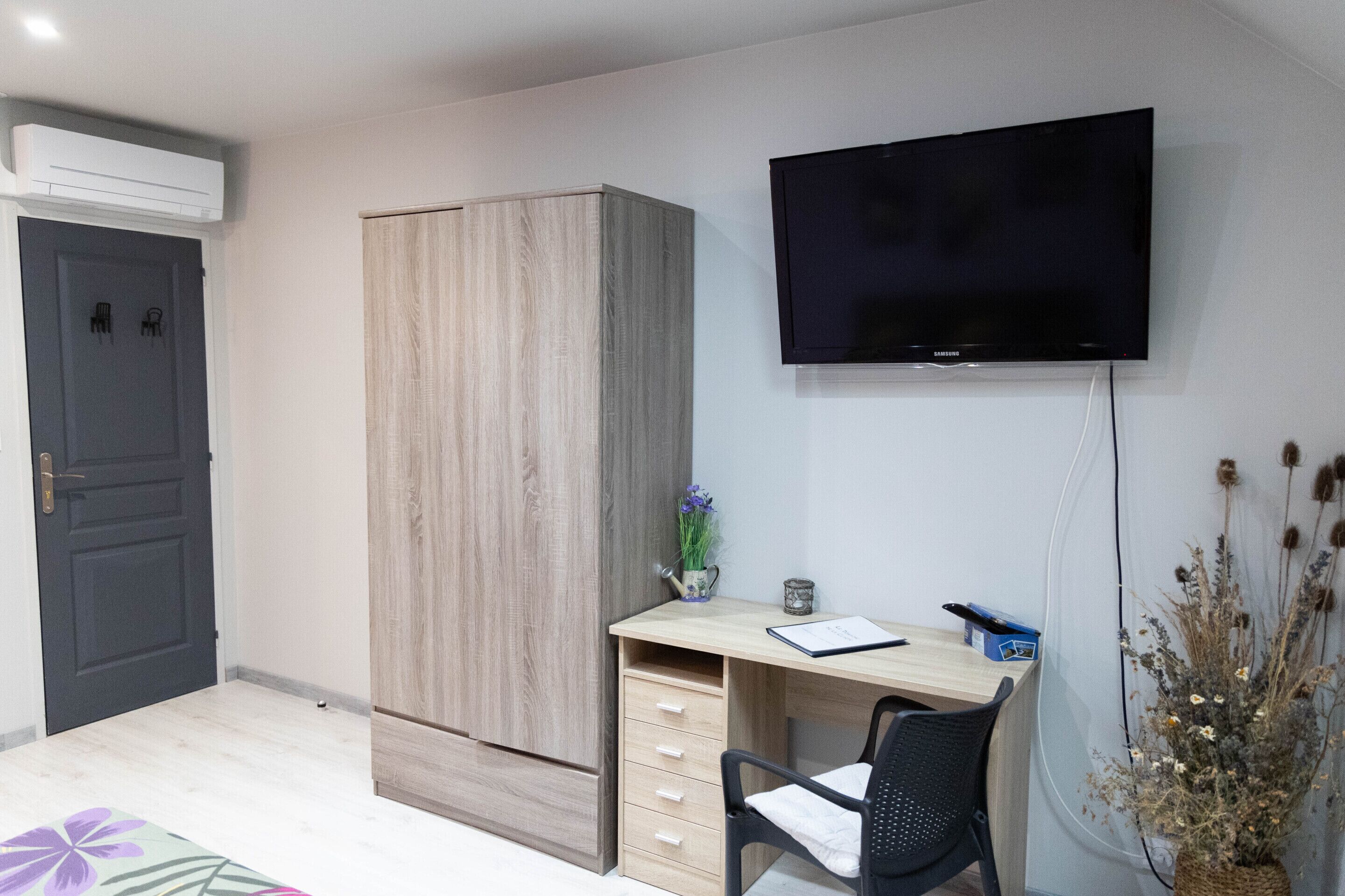 A room with a wooden wardrobe, a desk with a chair, a flat-screen TV, and a vase with dried flowers.