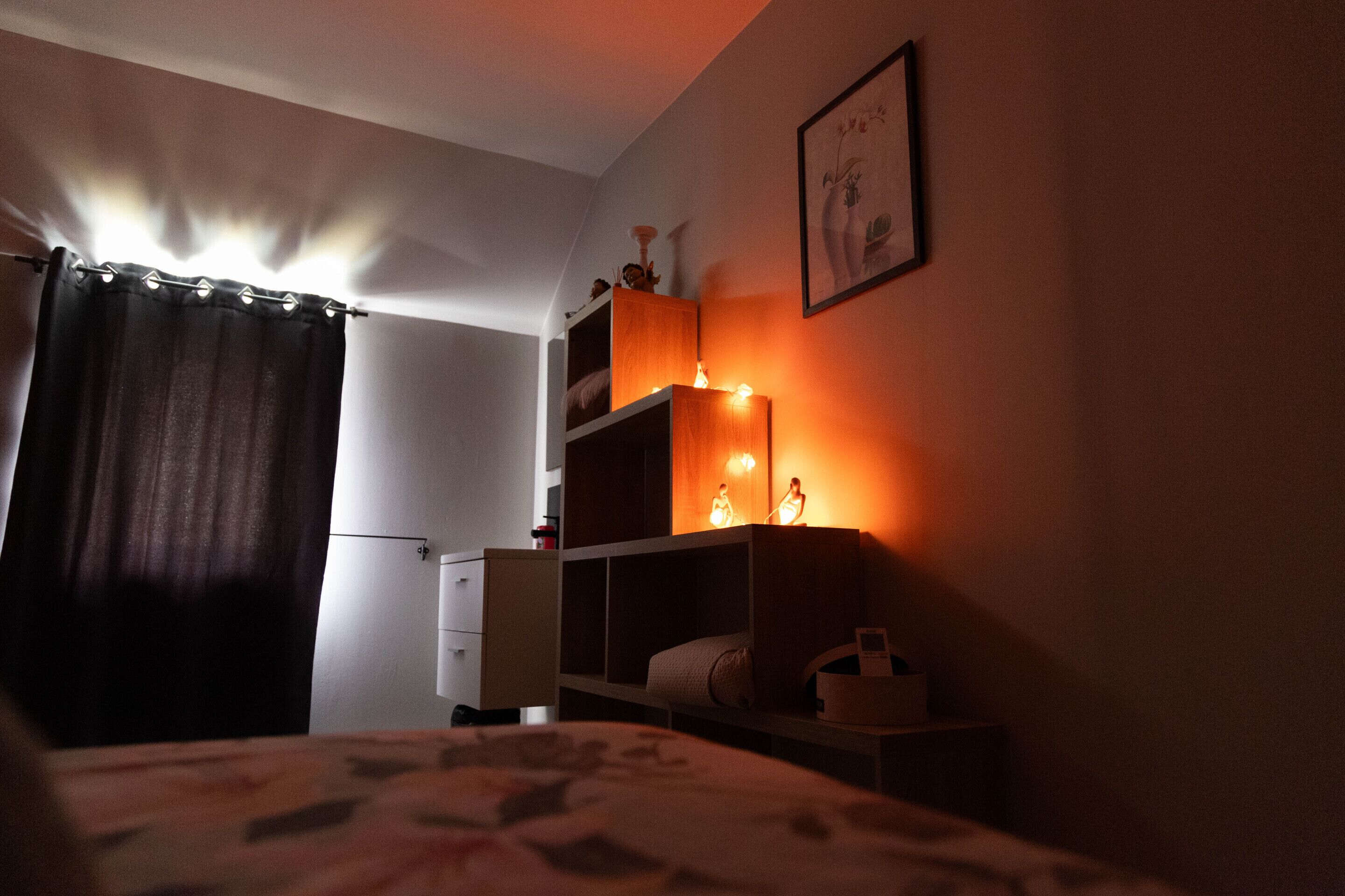 Un dormitorio con poca luz, una cama, un aparador y una estantería con adornos.