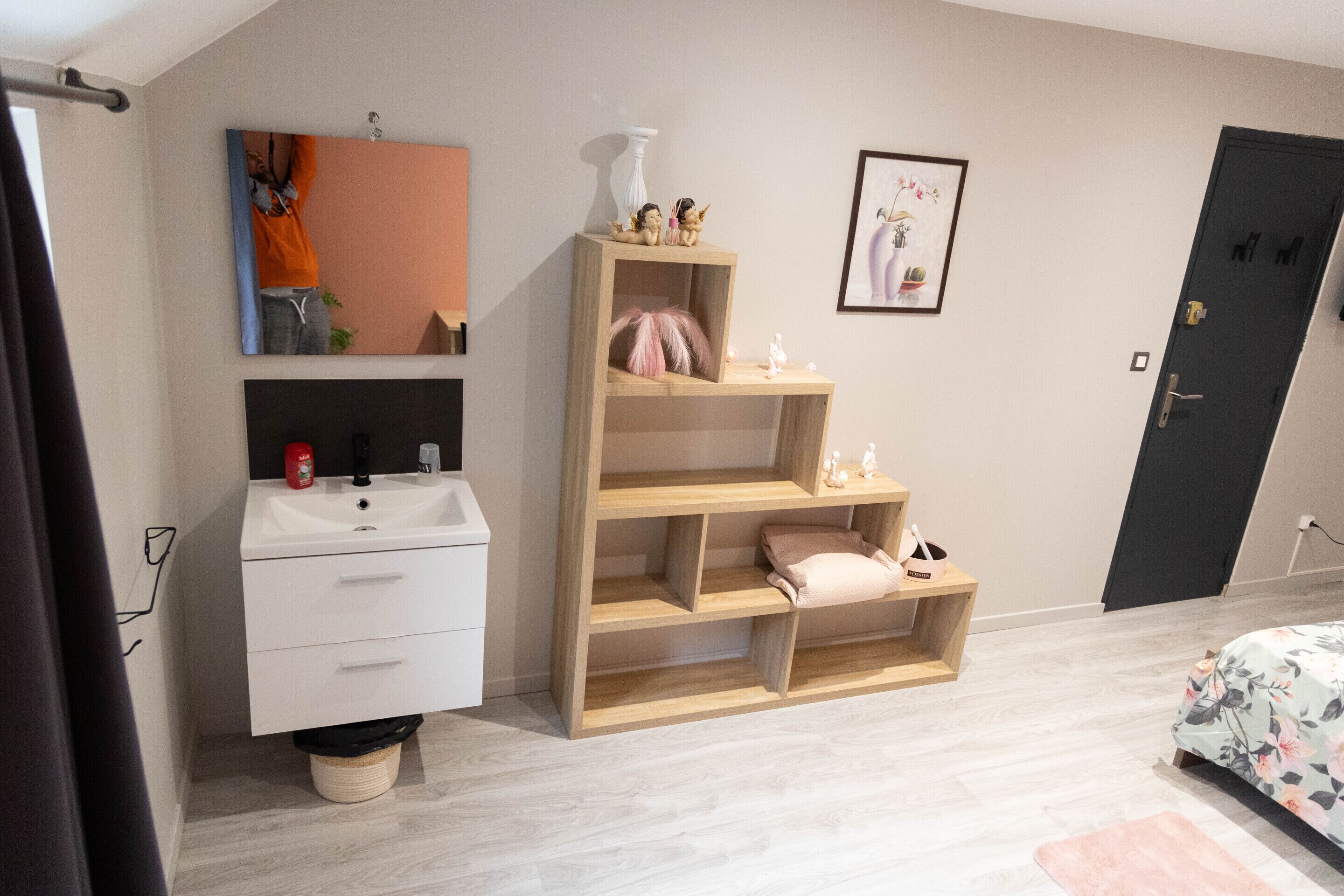 A bedroom with a wooden shelf unit, a white bedside table, a bed with floral bedding, and a door with a handle.