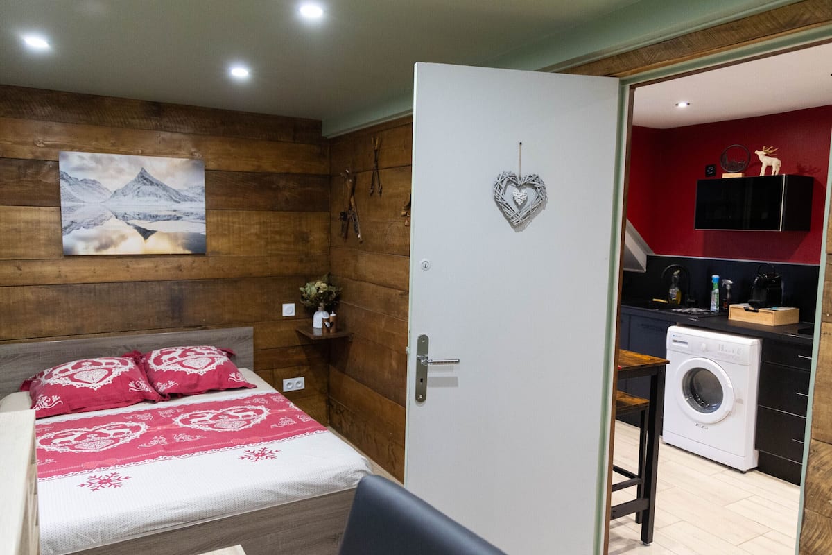 Ein Schlafzimmer mit einem Bett, einer Holzwand, einem Gemälde und einer Tür zu einem Küchenbereich mit einer Waschmaschine.