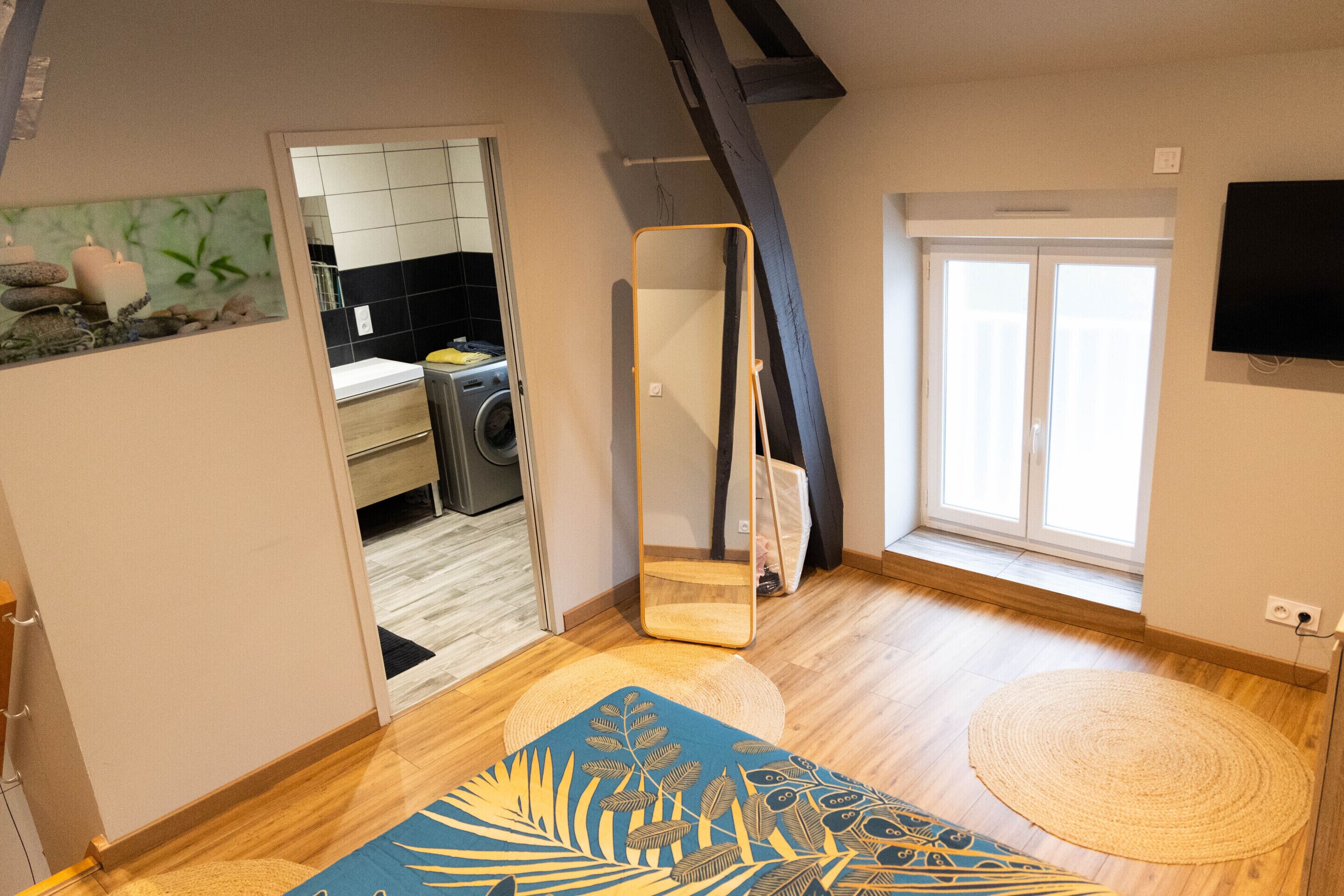 Quarto com um espelho grande, um tapete azul estampado, um tapete tecido, assoalho de madeira e uma TV fixada na parede.