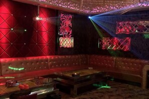 Una habitación con poca luz, una pared iluminada en rojo, una zona de estar con cojines y una mesa con una bebida.