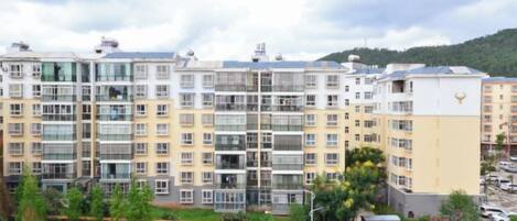 A multi-story residential building with balconies, a parking area, and a hilly landscape in the background.
